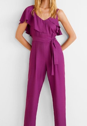 Kvinde iført en lilla ærmeløs jumpsuit med flæsedetalje på den ene skulder og bælte bundet i taljen, stående mod en hvid baggrund.