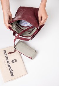 Bordeaux lederen tas met een gestreepte binnenkant, toont een groene pouch en een beige stoffen tas met een logo. Voorzien van een verstelbare schouderband en ritsen.