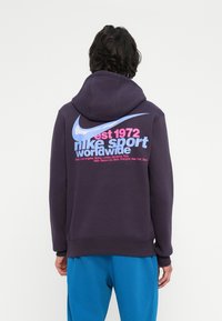 Pessoa vestindo um capuz roxo escuro com um grande swoosh azul da Nike e o texto em rosa e azul claro "est 1972 nike sport worldwide" nas costas.