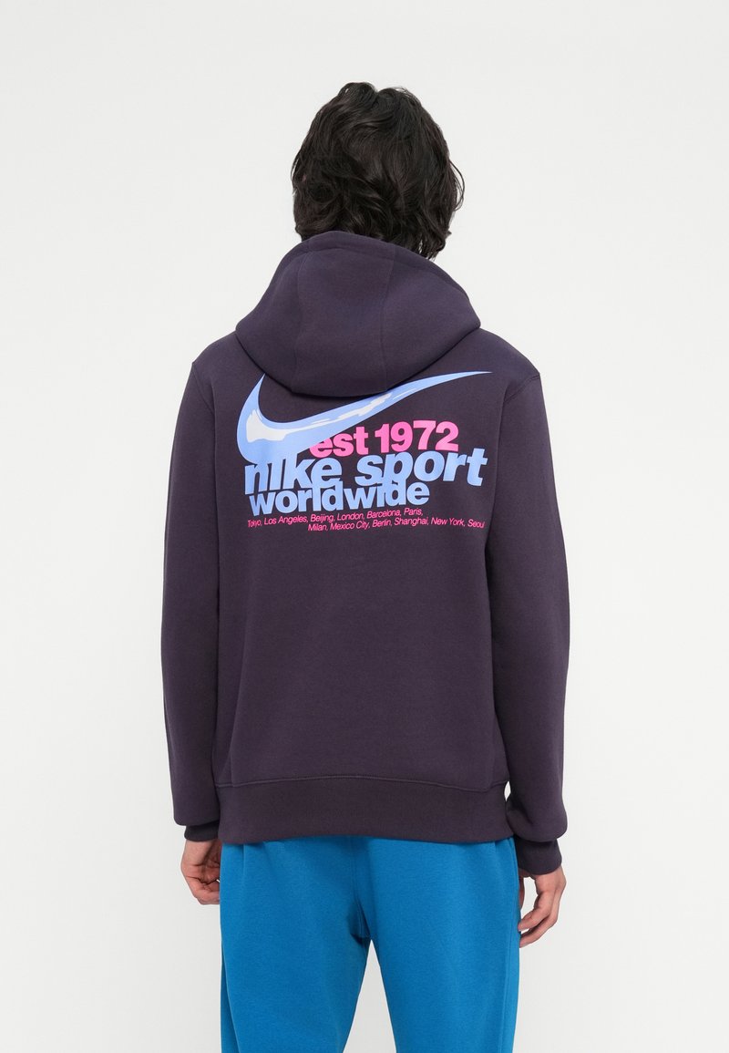 Pessoa vestindo um capuz roxo escuro com um grande swoosh azul da Nike e o texto em rosa e azul claro "est 1972 nike sport worldwide" nas costas.