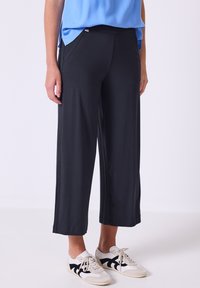 Pantaloni neri a gamba larga, corti, realizzati in tessuto morbido, con tasche laterali e una cintura comoda, abbinati a sneakers bianche.