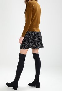 Brauner Pullover mit Rüschendetail, kombiniert mit einem schwarzen Minirock mit Punkten. Das Model trägt schwarze Overknee-Stiefel mit Blockabsätzen.