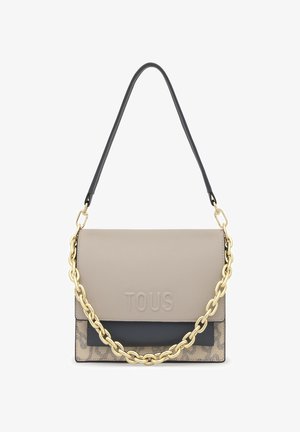 Bolso rectangular beige y negro con el logo "TOUS" en relieve, detalle de cadena dorada y asa de hombro negra sobre un fondo blanco.