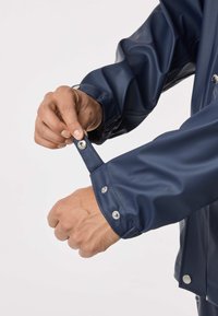 Marineblauw waterdicht jack met een gladde textuur. De mouw heeft een drukknopsluiting en verstelbare manchetten. Voorzien van opvallende stiksels en een modern ontwerp.