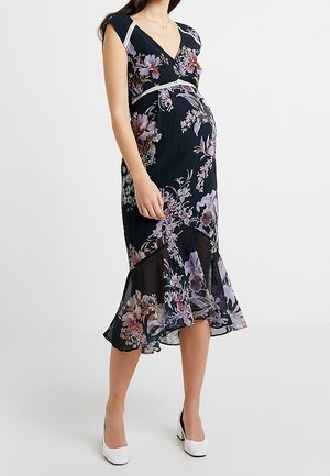 Donna incinta che indossa un abito midi nero a fiori con maniche a cappuccio, scollo a V e tacchi bianchi, in piedi davanti a uno sfondo chiaro uniforme.