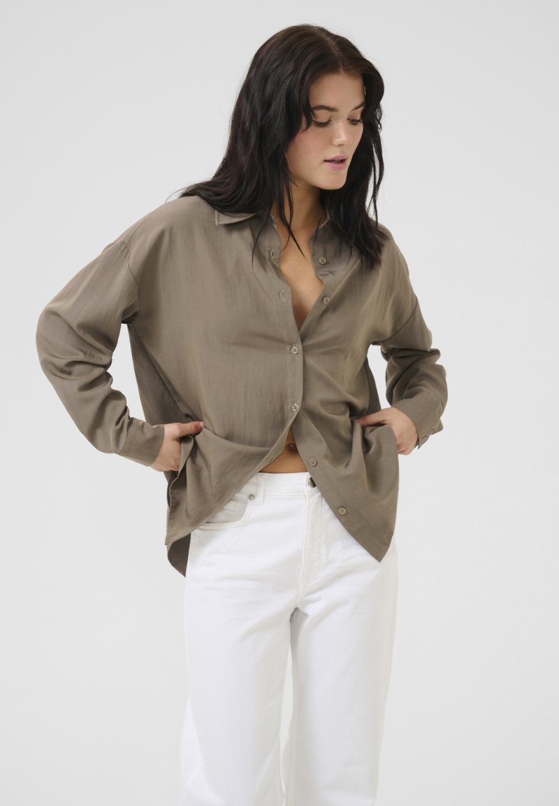 Brown button-up shirt with a relaxed fit, long sleeves, and a collared neckline, paired with white jeans. Smooth texture, minimal design.