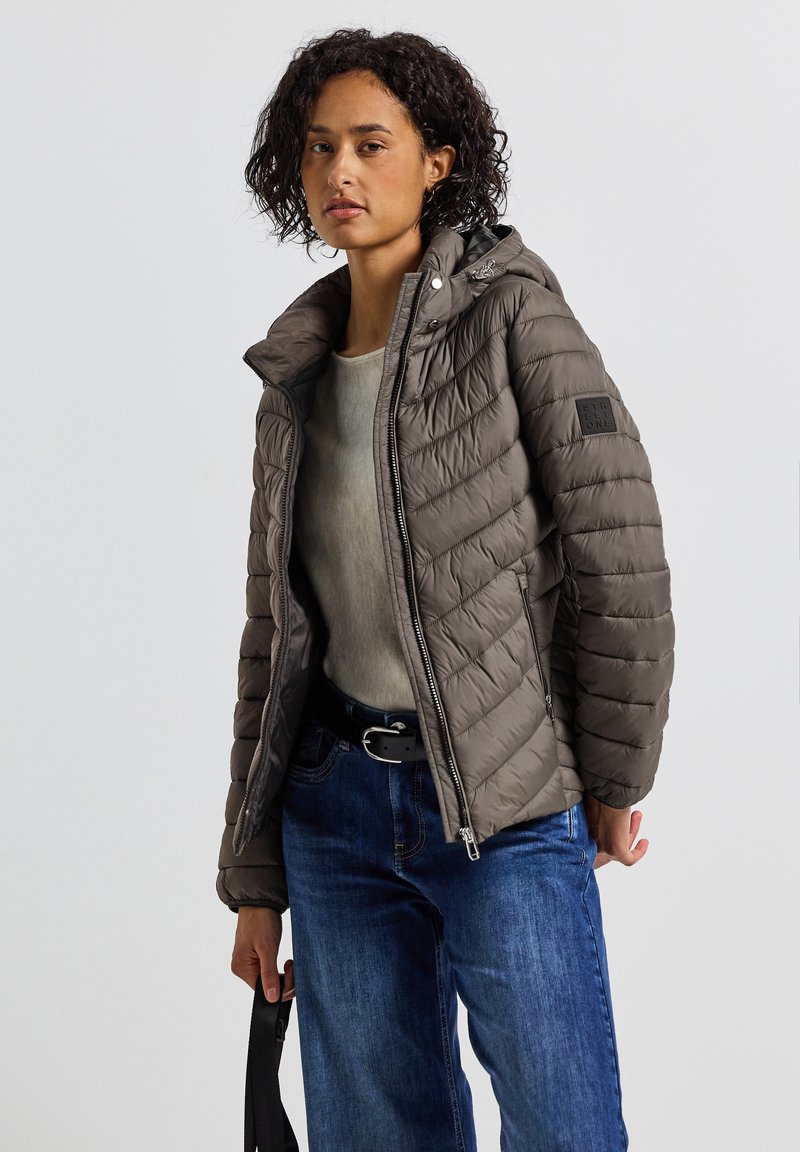 Veste rembourrée marron foncé avec fermeture éclair, capuche ajustable et poches latérales. Portée par-dessus un pull léger et un jean bleu. Design matelassé texturé.