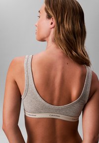 Sujeción deportiva de algodón gris con tirantes anchos, con un diseño minimalista y una banda de logo beige en la parte inferior.