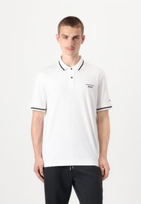 Balta polo marškinių, juodu apykakle ir rankovėmis, su dviem mygtukais ir siuvinėtų "Porsche BOSS" logotipu kairėje krūtinės pusėje.
