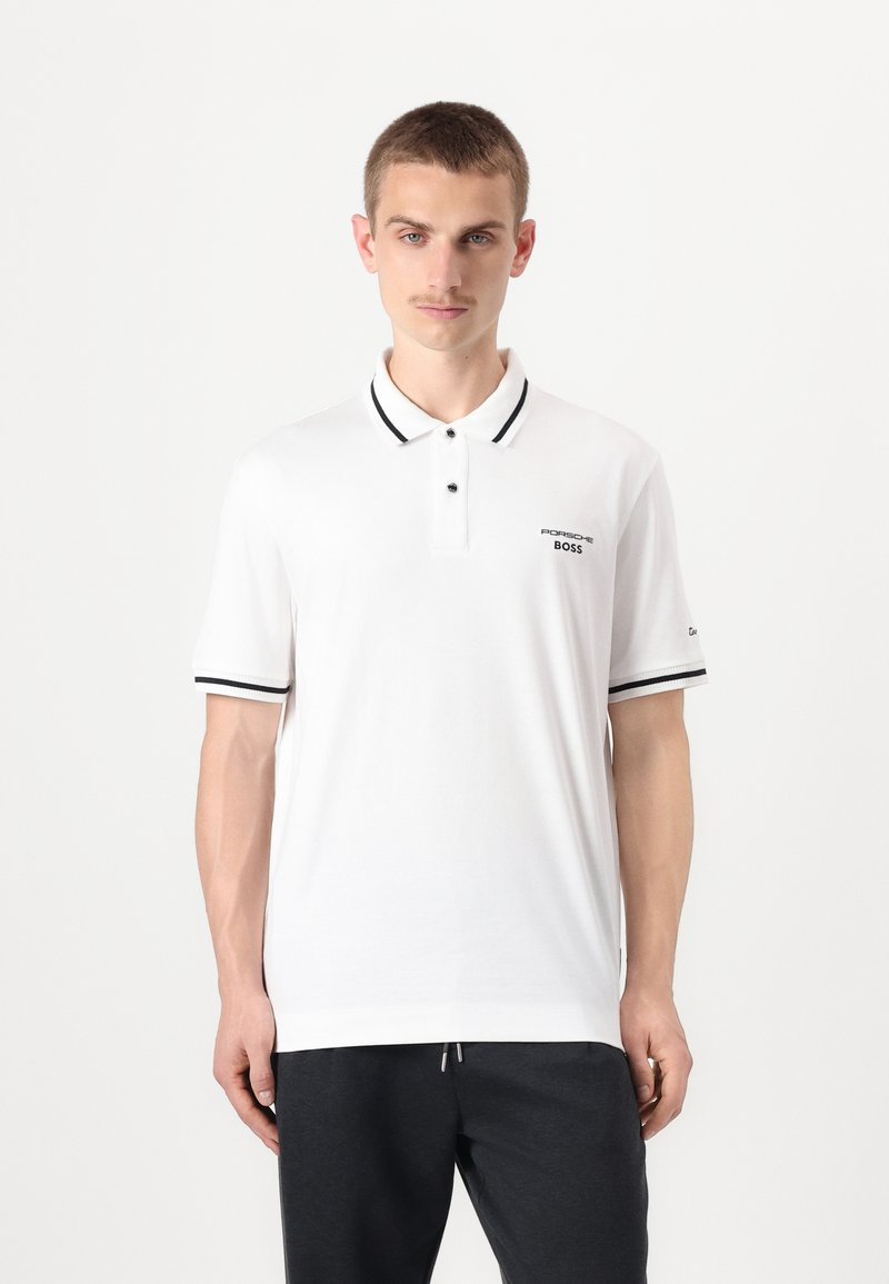Balta polo marškinių, juodu apykakle ir rankovėmis, su dviem mygtukais ir siuvinėtų "Porsche BOSS" logotipu kairėje krūtinės pusėje.