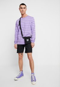 Fialovo prúžkovaná košeľa s dlhým rukávom, čierne šortky a fialové tenisky s vysokým strihom. Pri páse je zavesená malá čierna crossbody taška.