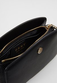 Portafoglio in pelle nera con texture, dotato di cerniera dorata e chiusura a scatto, parzialmente aperto che mostra l'interno nero e l'etichetta DKNY New York.