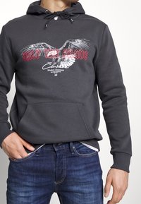 Sudadera gris con un bolsillo frontal, que presenta un gráfico de un águila y "SOLO LOS VALIENTES" en texto rojo. Material de algodón con capucha ajustable.