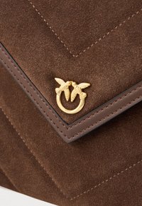Hnědá semišová kabelka s designem ve tvaru chevron, zlatými prvky ve tvaru ptáčka a jemným prošíváním podél okrajů.