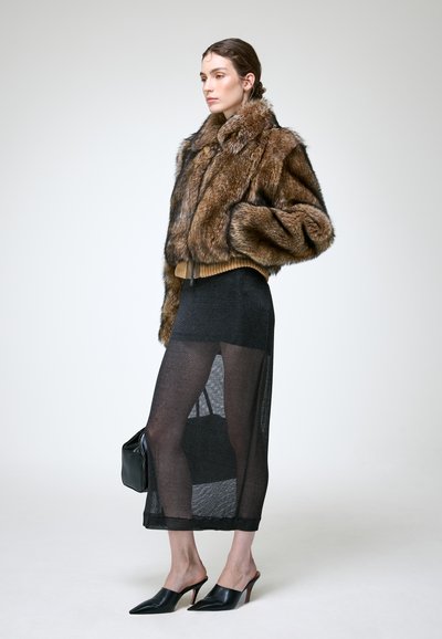 Femme portant une veste en fourrure marron, une jupe noire transparente, des mules noires à talons hauts, tenant un sac à main noir sur un fond uni.