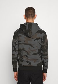 Hooded sweatshirt i kamouflage-mönster med mörkgröna, grå och svarta nyanser. Materialet är mjukt med ribbade muddar och nederkant.