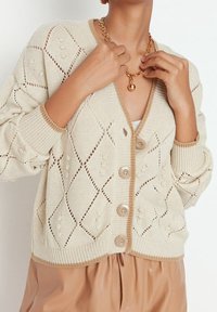Beige knitted cardigan with a diamond pattern, features pearl accents, a V-neckline, and five buttons down the front. Ribbed cuffs and hem.