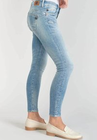 Vaqueros ajustados azul claro hechos de denim con un lavado desgastado, que cuentan con bolsillos traseros y un parche con el logo en la cintura. Combinados con mocasines crema.