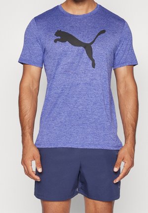 Mężczyzna w fioletowej koszulce z krótkim rękawem z czarnym logo Puma oraz granatowych sportowych szortach, stojący na jednolitym tle.