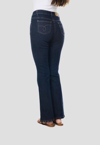 Donkerblauwe uitlopende spijkerbroek van denim, voorzien van zakken met decoratieve stiksels. De zoom is lichtelijk rafelig. Draagt bijpassende bruine sandalen.