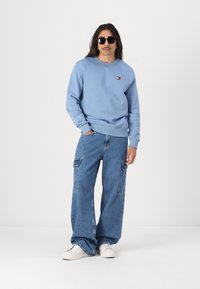 Felpa azzurra chiaro con scollo rotondo e piccolo logo, abbinata a pantaloni cargo blu in denim oversize e sneakers bianche. Occhiali da sole neri indossati.