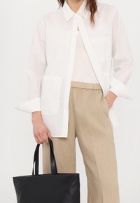 Chemise blanche boutonnée avec col et poche poitrine, associée à un pantalon en lin beige. Sac cabas noir à longues anses tenu à la main.