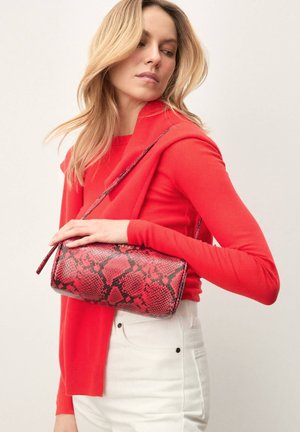 Sac à main rouge à imprimé serpent avec une forme arrondie, en cuir texturé, et une fine bandoulière portée en travers du corps. Le mannequin porte un haut rouge et un pantalon blanc.
