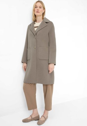 Frau trägt einen taupefarbenen knielangen Mantel mit großen Taschen, beige gekürzte Hose und passende flache Schuhe, steht vor weißem Hintergrund.