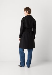 Černý trenčkot s opaskem v pase, dlouhými rukávy a širokým límcem, nošený přes volné modré džíny. Minimalistický design a hladká textura.