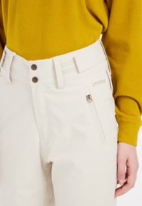 Bež snježne hlače s glatkom teksturom, dva smeđa dugmeta, bočnim džepom na zip, i logotipom blizu pojasa. Uparen s žutom puloverom.