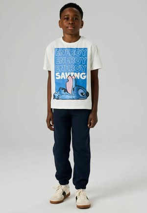 Camiseta de algodón blanca con un estampado de un personaje de dibujo animado en azul y rosa, y texto en negrita que dice "AHORRO DE ENERGÍA", combinada con pantalones deportivos azul marino y zapatillas.