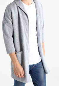 Gray knitted cardigan with a shawl collar, three-quarter sleeves, and front pockets, displayed over a white t-shirt and blue jeans.