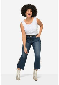 Top blanco sin mangas con botones y un bolsillo, combinado con jeans cortos azul oscuro que tienen acentos de botones en los dobladillos y botas de tobillo plateadas.
