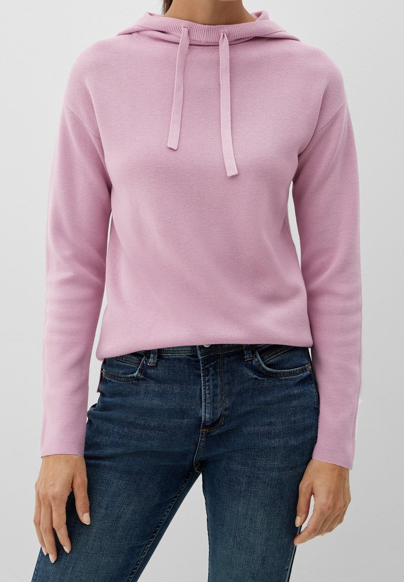 Femme portant un sweat à capuche rose clair avec cordons et un jean ajusté bleu foncé, debout devant un fond clair uni.