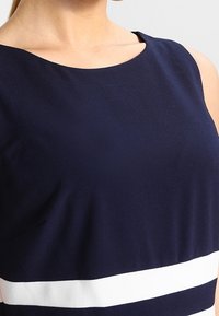 Námořnická modrá tílko bez rukávů s kulatým výstřihem. Obsahuje bílý vodorovný pruh blízko lemu. Hladká textilie.