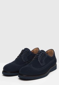 Tyrkysové semišové brogue boty mají design s křídlovými špičkami, perforované detaily a kontrastní černou gumovou podrážku. Šněrování s šesti očky.