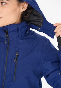 Dunkelblaue Jacke mit hohem Kragen, Reißverschluss vorne, verstellbare Kapuze und schwarer Innenfutter. Mit Reißverschlüssen und Klettverschluss-Akzenten.