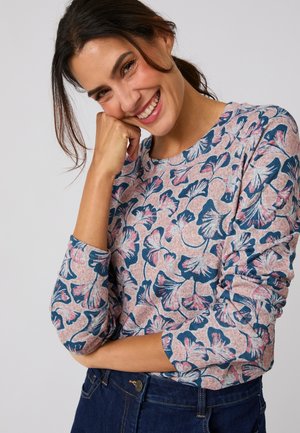 Glimlachende vrouw met donker haar in een paardenstaart, draagt een top met lange mouwen en bloemenpatroon en donkere blauwe spijkerbroek op een effen achtergrond.