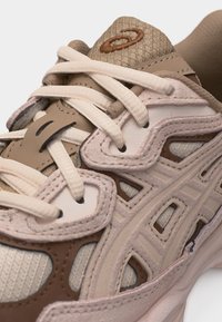 Beige and brown athletic shoe with thick white laces, textured fabric tongue, and layered synthetic overlays for support and style.