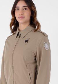 Giacca leggera beige con colletto strutturato, chiusura con zip, tasche laterali e una patch con logo sulla spalla sinistra. Texture liscia.