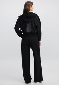 Sudadera negra con capucha y un panel trasero brillante, combinada con pantalones negros de pierna ancha. Textura suave, ajuste relajado, diseño minimalista.