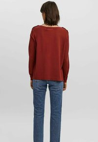 Maglione color ruggine con vestibilità ampia, scollatura asimmetrica e bottoni decorativi sulle spalle, abbinato a jeans a gamba dritta blu.