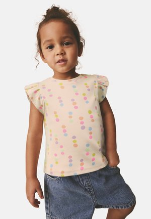 Unge barn med krøllet hår iført en cremefarvet skjorte med farverige prikker og denimshorts, poserer mod en ensfarvet baggrund.