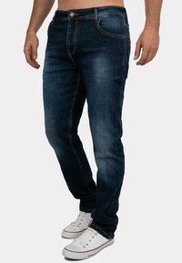 Jeans in denim blu scuro con vestibilità slim, caratterizzati da cuciture arancioni, cinque tasche e chiusura con bottone. Indossati con sneakers bianche.
