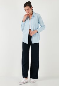Camicia button-up azzurro chiaro con maniche lunghe, vestibilità leggermente over-size, tessuto testurizzato, abbinata a pantaloni larghi scuri e sneakers bianche.