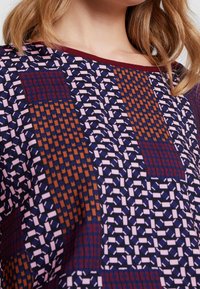 Blusa con estampado que presenta una mezcla de diseños geométricos en colores marino, rosa y óxido, con cuello redondeado y textura de tela suave.