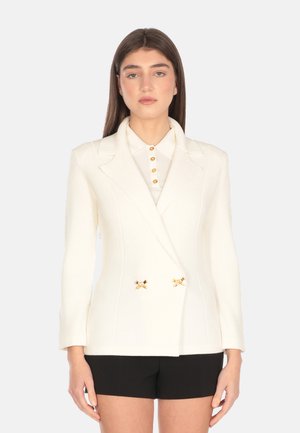 Blazer bianco a costine con design doppiopetto, revers, dettagli in metallo dorato e maniche lunghe, abbinato a una camicia con colletto.