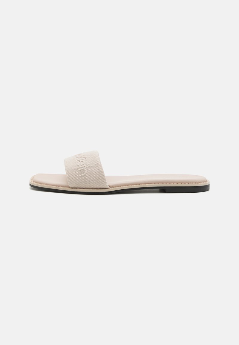 Beige slidesandal med en bred stofrem, glat fodseng og en slank sort sål. Remmen har præget branding.