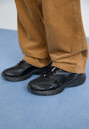 Osoba nosząca czarne buty sportowe i luźne brązowe spodnie sztruksowe stojąca na niebieskim dywanowym podłożu.
