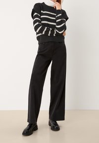 Schwarze weit geschnittene Hose kombiniert mit einem gestreiften Dunkelgrau- und Cremefarbenen Pullover. Der Pullover hat eine gestrickte Textur und gerippte Bündchen.